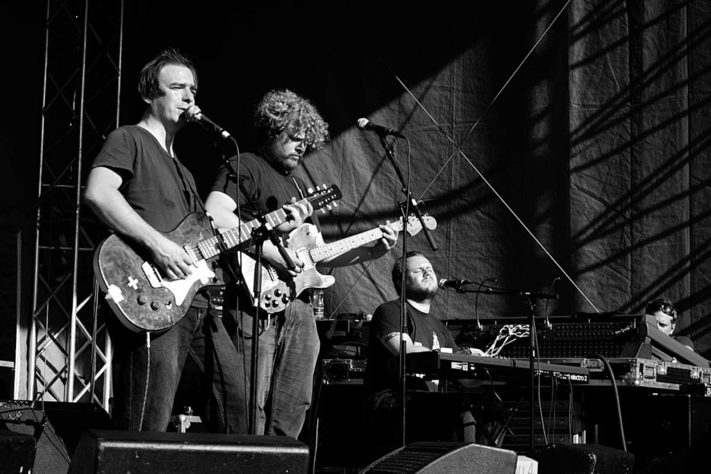 Black and white photograph of Jason Molina on stage playing guitar with various band members on their instruments.