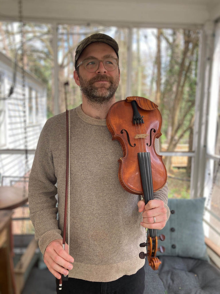 A man standing on a screened in porch with the side of the house and trees/yard showing behind him. He is bearded and wearing glasses,a tan sweater, a baseball cap, and dark pants. He holds in fiddle in one hand and a bow in the other.