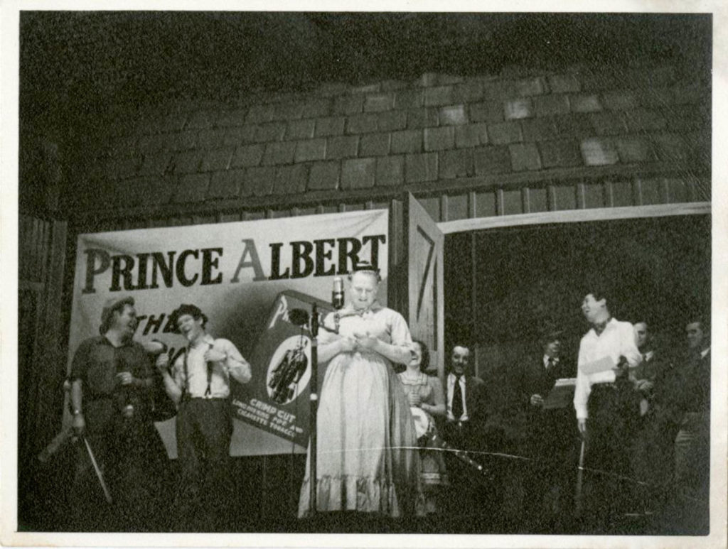 Several musicians and background people on the stage with a Prince Albert tobacco advertisement hung on the wall behind them. The Duke of Paducah is center stage, dressed as a woman.