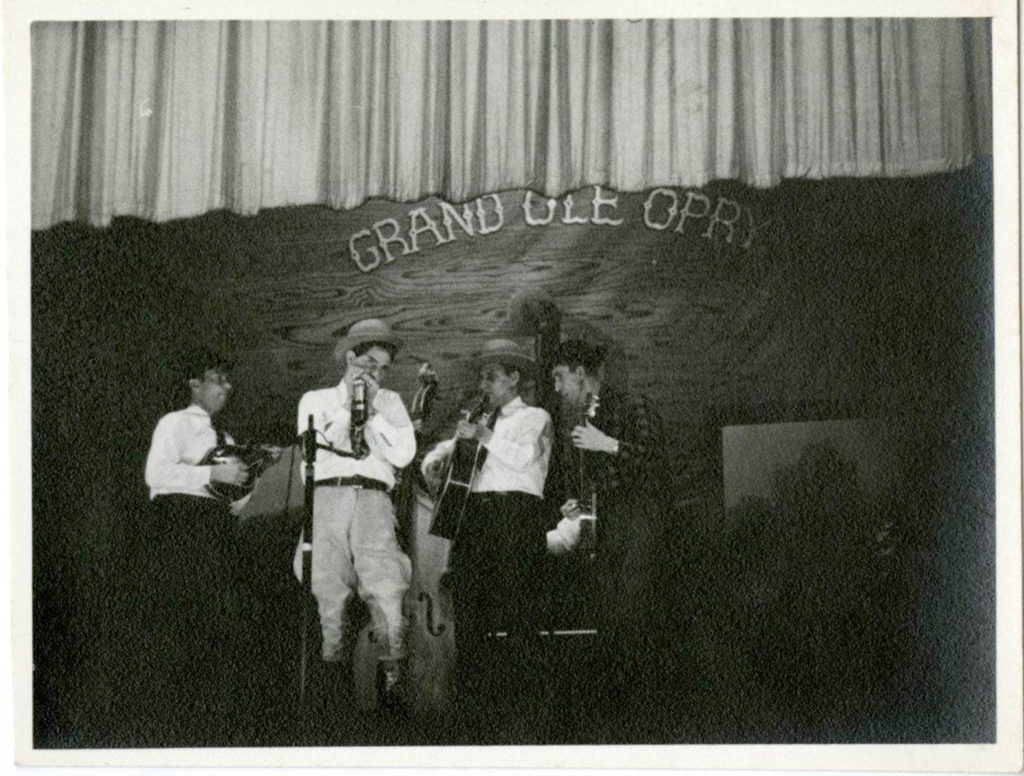 Four musicians, all wearing hats of various styles, on the Grand Ole Opry stage, gathered round the central mic. Far left: mandolin player, near left: Curly Bradshaw playing harmonica, near right: Bill Monroe playing guitar, and far right: Stringbean playing banjo.