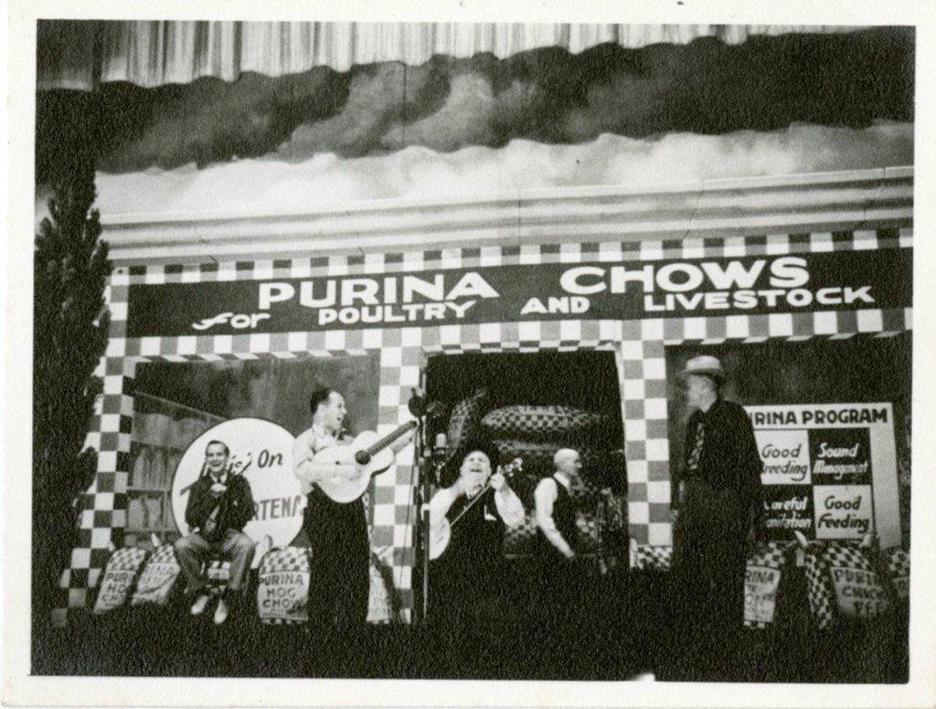 The Opry stage decorated with a backdrop for Purina Chows for Poultry and Livestock. Four musicians, plus a man in the background, are seen, including Dorris Macon on guitar and Uncle Dave Macon on banjo.