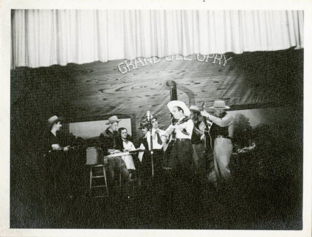 Eight musicians and background people on the Opry stage, playing a variety of instruments. Zeke Clements is front and center playing the guitar at the mic.