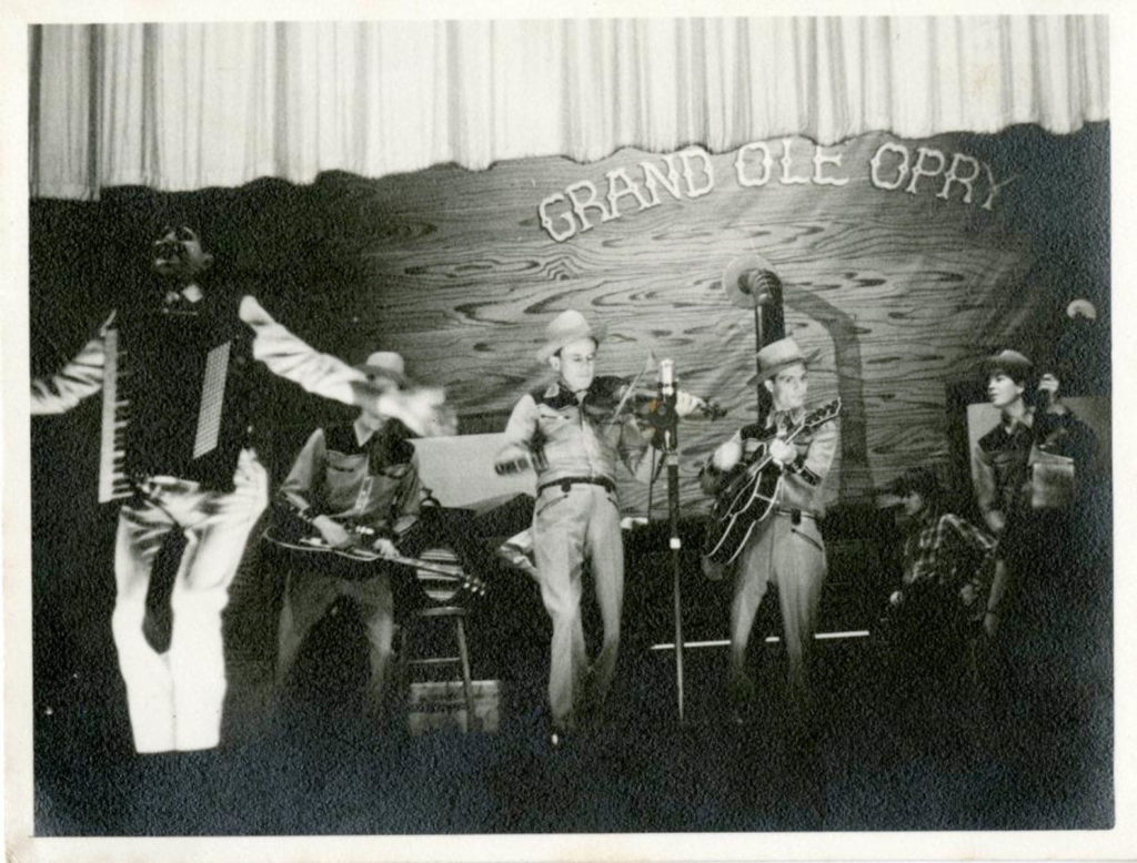 Pee Wee King is to the far left with his accordion and playing to the audience. He is backed by four musicians in matching outfits and playing a variety of instruments.