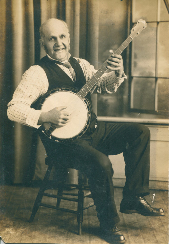 Uncle Dave Macon in a shirt, vest and trousers playing the banjo sitting down.
