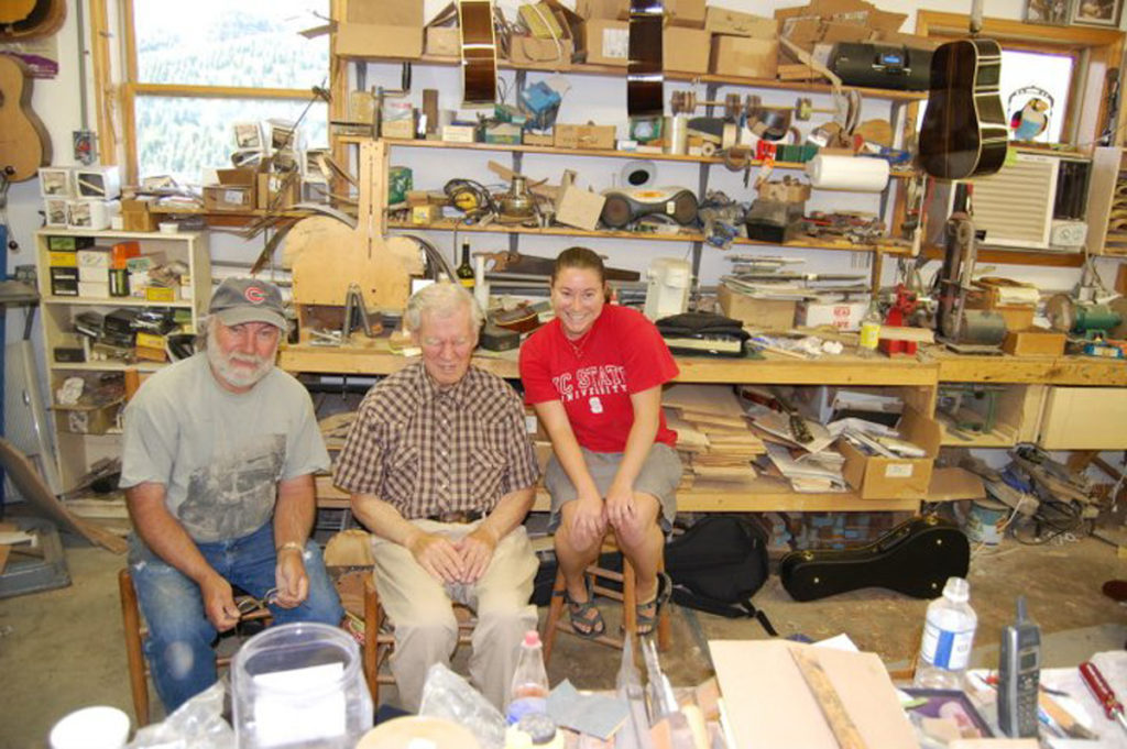 Wayne Henderson, Doc Watson, and Jayne Henderson posing for the camera in the Henderson workshop. Tools, instruments, and wood litter the background of the photograph.