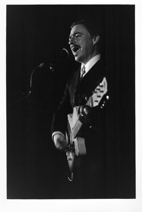 Black and white close-up of Jason Molina wearing a dark suit and with his guitar on stage.