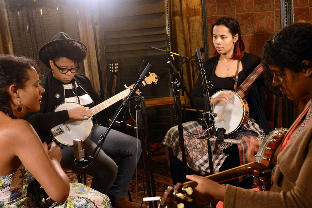 The four musicians sit in a small circle in studio, playing their banjos with mics and other equipment around them.