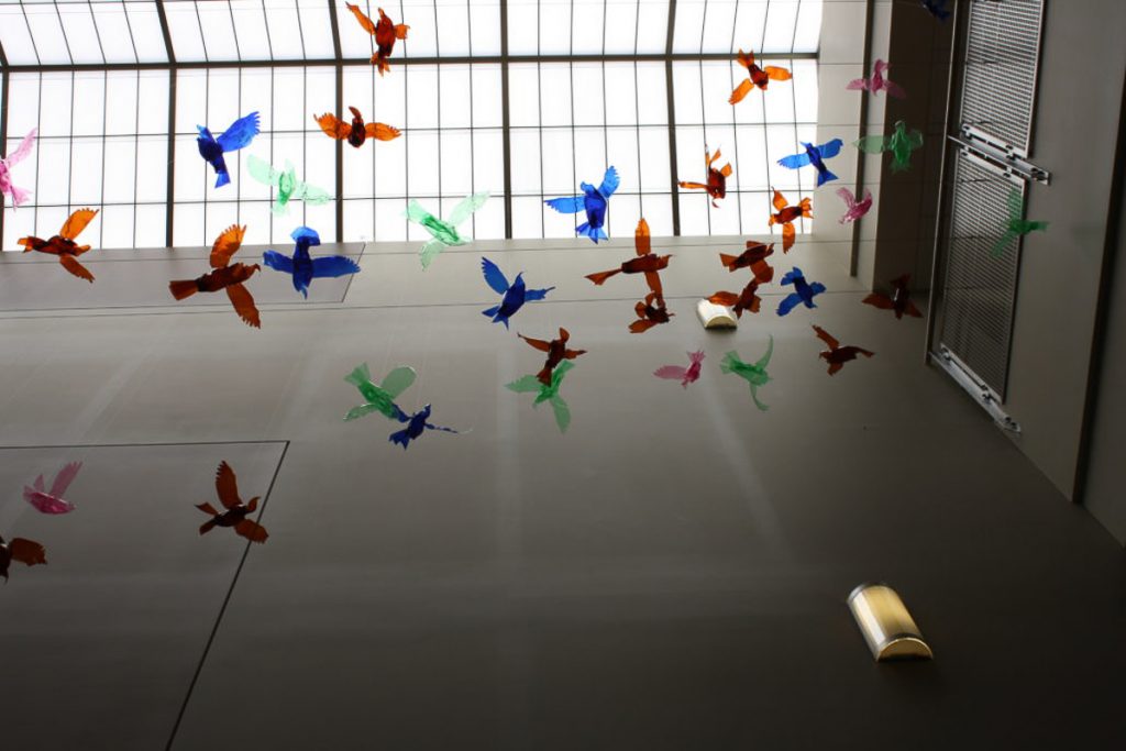 Looking up towards the glass ceiling of the building, the photo shows several colorful birds (blue, green, pink, orange, and red) in flight made of plastic bottles.