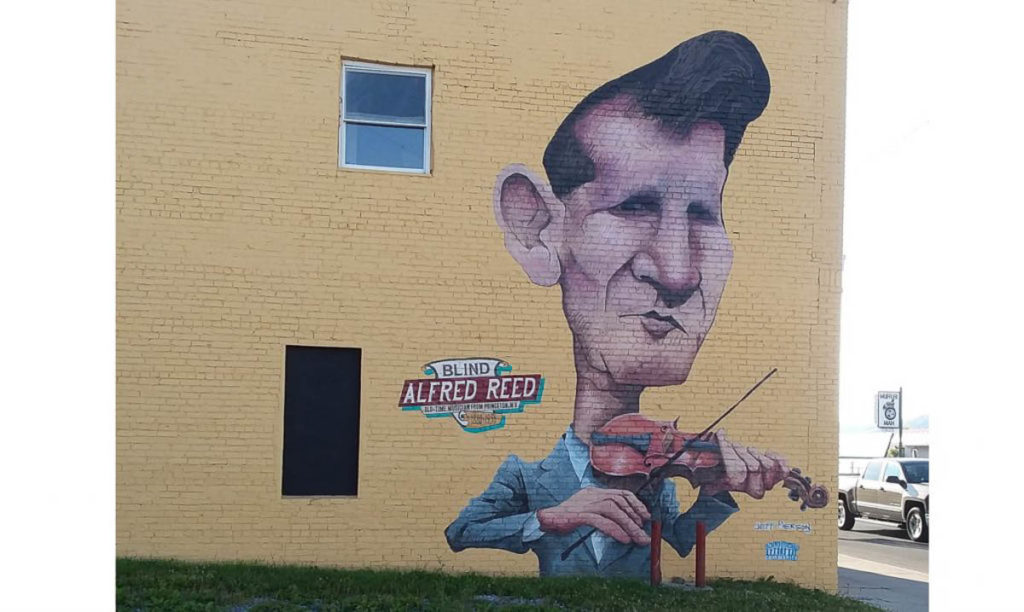 An image of a large cream-colored wall with a cariacature-style portrait of Blind Alfred Reed to the right on the wall. His dark hair is in a pompadour style, his ear is unusually big, and he holds his fiddle to his chin. He wears a greyish blue suit; the words "Blind Alfred Reed" are written to the left of the portrait.