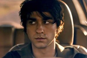 Actor Alex Wolff in the driver's seat of his car looking pensively into the rear view mirror.