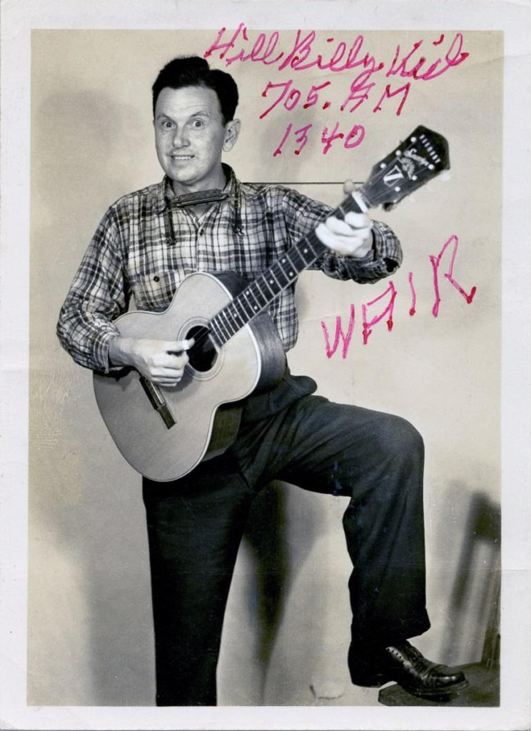 Photograph showing man in check shirt with dark pants, one foot up on a chair and holding a guitar. He has a harmonica around his neck. Written on the photograph is "Hillbilly Kid / 705.AM 1340, / WAIR