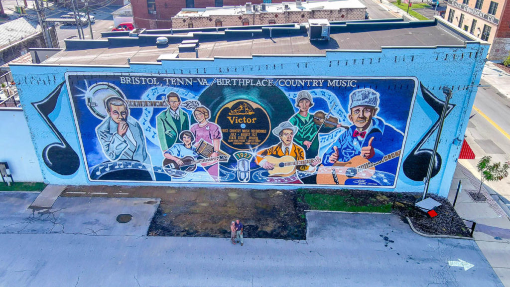 This aerial view shows the side of a building painted with a wall-sized mural. Two music notes bookend the central painting that bears these words at the top "Bristol, Tenn-VA / Birthplace of Country Music." The central painting show several people or groups of people in a graphic/realistic style, from left to right: Ralph Peer (in a grey suit and with grey hair), The Carter Family (A.P. Carter wearing a greenosh suit stands beside of Sara in a pink dress and holding an autoharp with Maybelle sat in front of them in a blue dress and playing her guitar), Ernest and Hattie Stoneman (he is wearing a brownish suit and white cowboy hat and holds his guitar, she is standing behind him in a green dress with a bonnet-style hat on and playing the fiddle), and Jimmie Rodgers (dressed as a railway worker with engineer's cap, blue jacket, and red bow tie, he has his guitar and is holding two thumbs up). In the center is a Victor record and a microphone with 1927 on it.