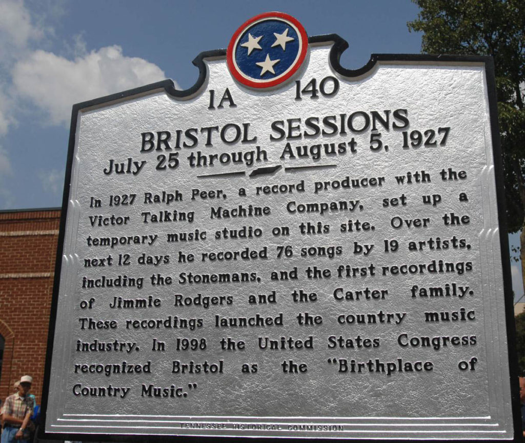 Large metal historic marker with the Tennessee symbol of three stars on a blue background with red border at the top. The words briefly describe the Bristol Sessions. A brick building can be seen in the background.