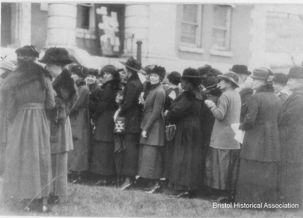A line of women crowd in front of a building. They are wearing early 20th century clotes, and one of the women looks out from the line and directly at the camera.