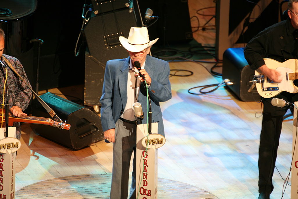  Charlie Louvin sings at a mic labeled WSM/Grand Ole Opry at center stage with other musicians and equipment around and behind him. He is wearing grey pants, a white shirt and Stetson, and a blue blazer.