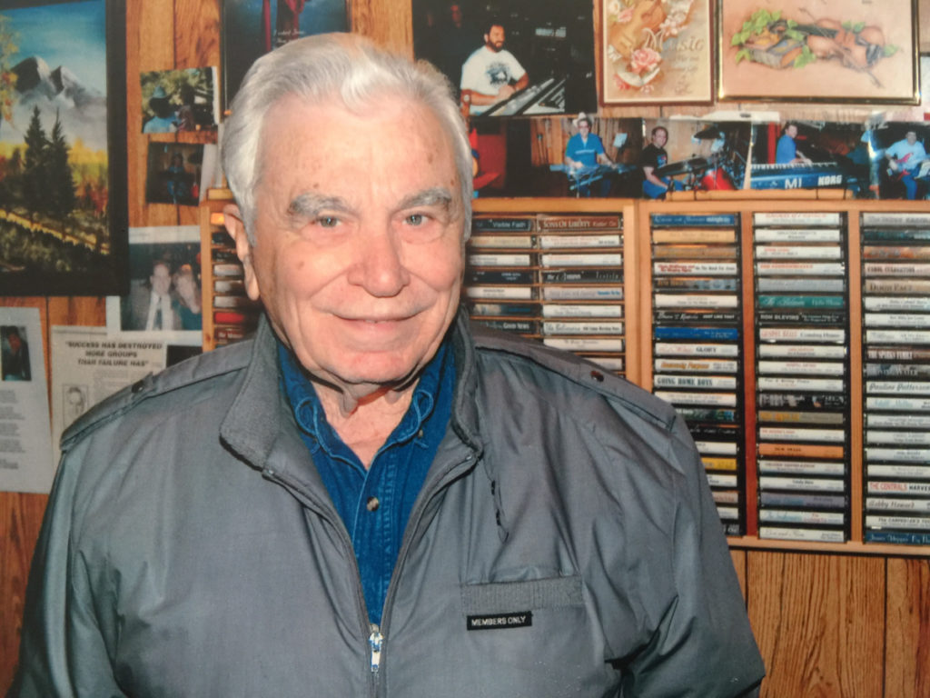 Charlie Maggard standing in front of a large display of cassette tapes with photographs of musicians who have recorded in Maggard Sound Studio on the wall.