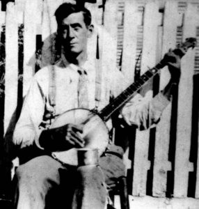 Black-and-white photograph of a dark-haired, clean-cut man seated in a chair in front of a white picket fence. He holds a banjo on his lap, ready to play.