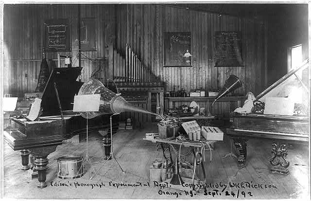 A black-and-white photograph of a large room filled with different musical instruments, including two pianos, a small drum, and what looks to be a small organ, along with several phonograph machines.