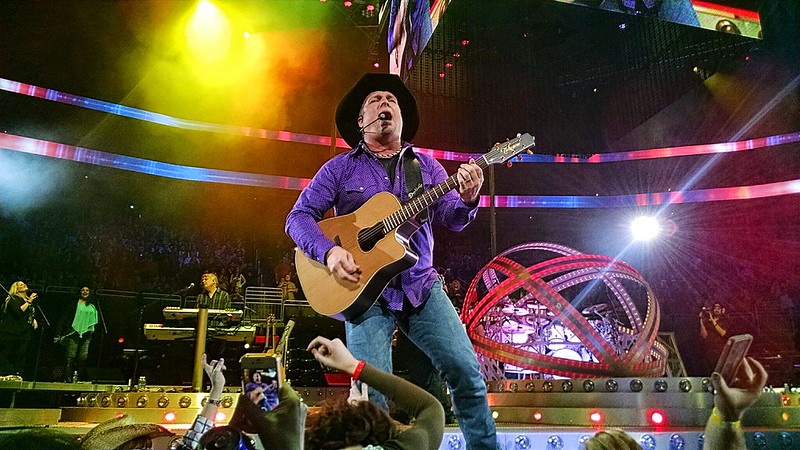 Garth Brooks on stage wearing jeans, a purple button-down shirt (untucked), and a black cowboy hat.