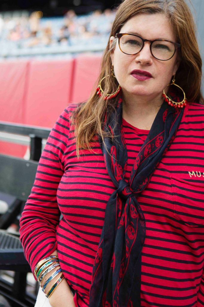 Picture of woman with long reddish brown hair, wearing glasses, a red and blue striped top, scarf, and big hoop earrings.