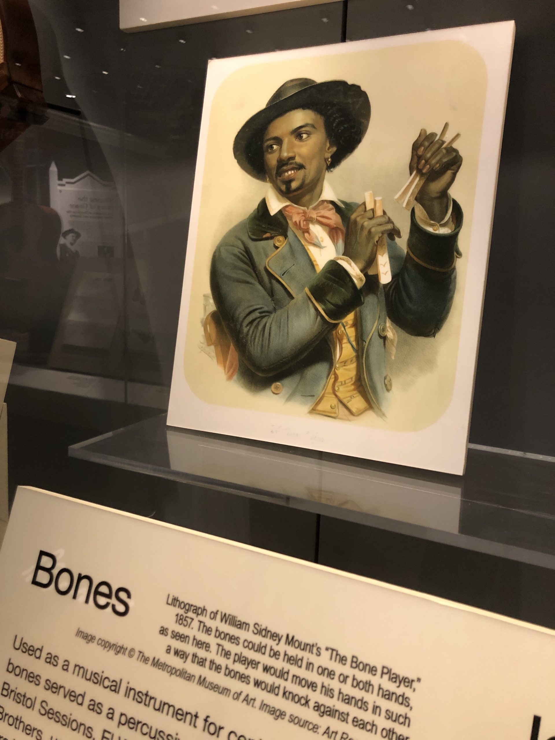An image of an exhibit case with William Sidney Mount's "The Bone Player" -- a black musician wearing a hat, jacket, waistcoat, and cravat-like tie, and holding two pairs of bones in his hands.