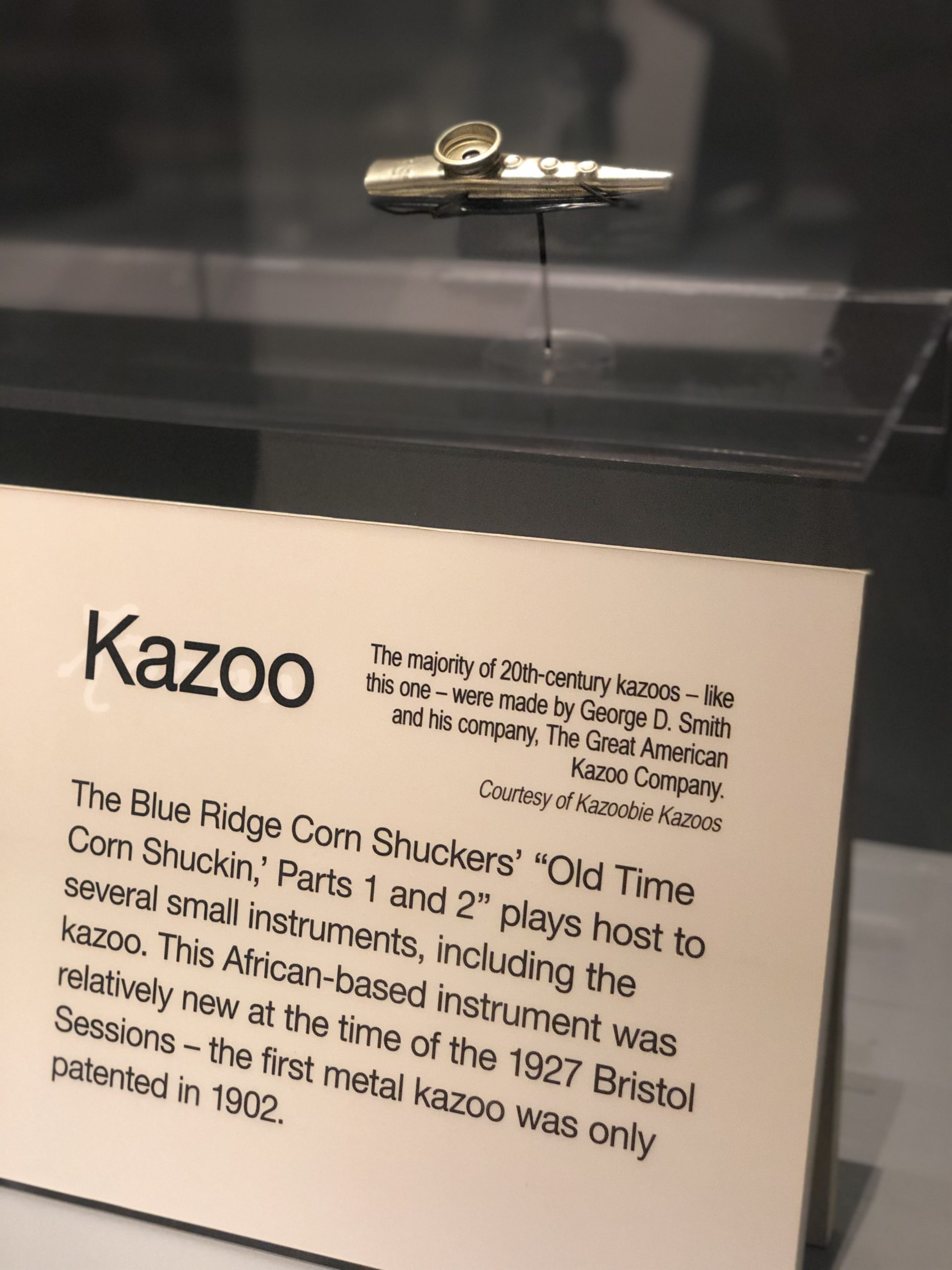A metal kazoo on a display stand within a glass case with an interpretive label in front of it with a brief text about the kazoo.