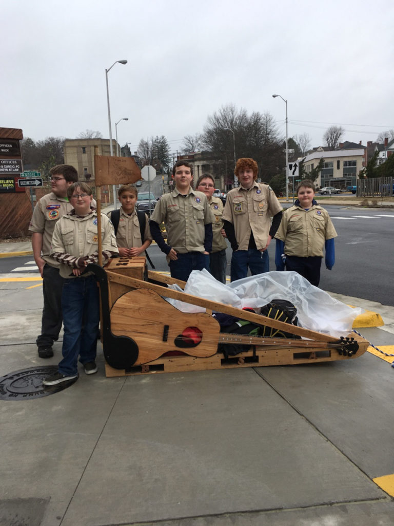 A picture of the Scouts posing with their sled in front of the museum -- the sled looks like a guitar laying on its side with a musical note as the handle.