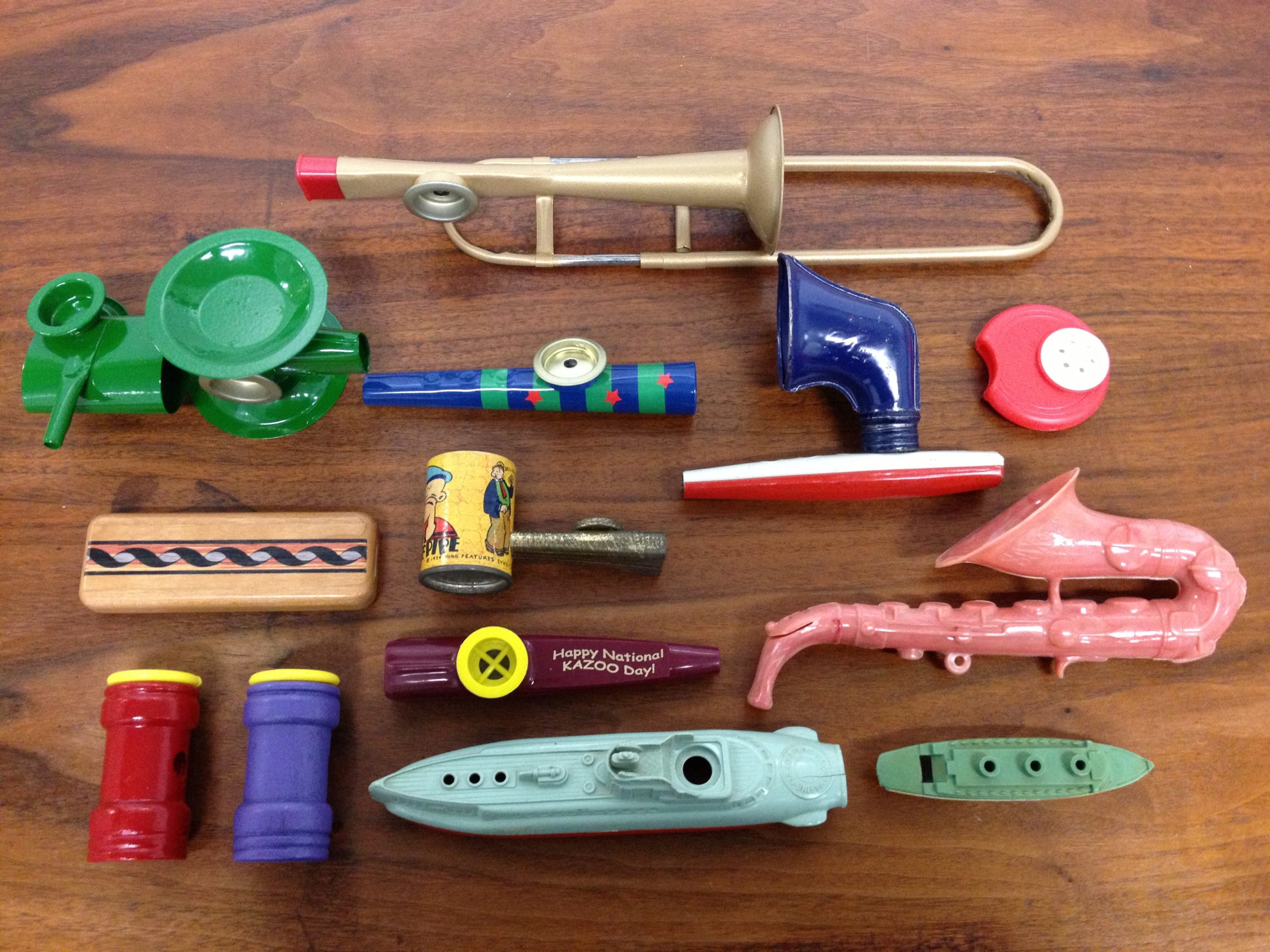 A variety of colorful plastic kazoos -- from common kazoo shapes to a pink saxophone shape to submarine/military ship shapes, to a trombone shape.