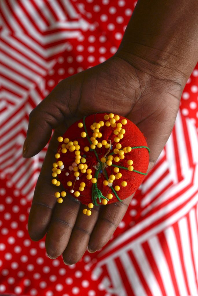 Close up of Sharon Tindall's hand holding a bright red pin cushion, filled with yellow head pins, over a red and white cloth.