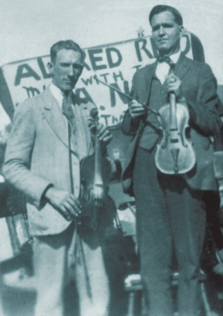 Photograph of Fred Pendleton holding a fiddle and Blind Alfred Reed holding a fiddle, standing in front of a sign advertising Reed's performance.