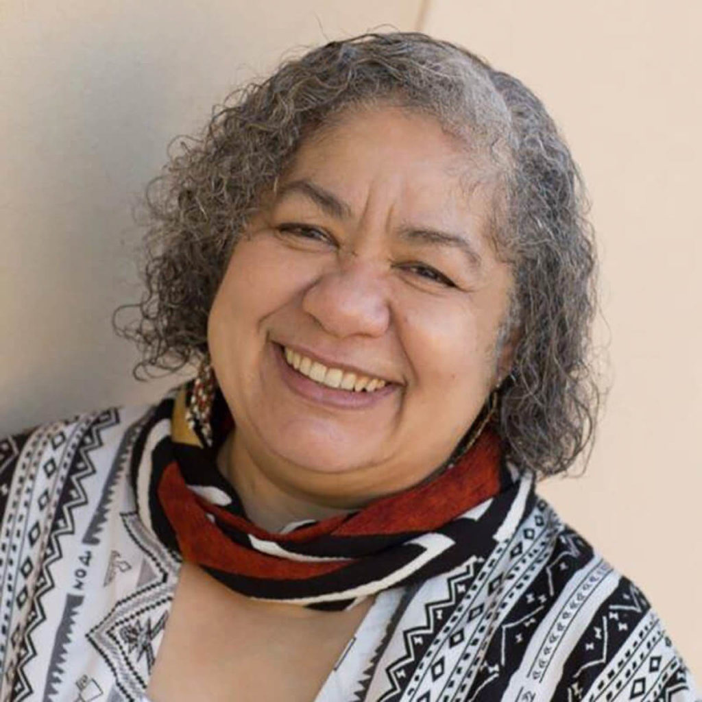 An image of a Black woman with grey, curly, chin-length hair. She is smiling widely. She wears a black and white patterned top with a red, black, and white scarf around her neck.
