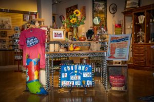 A general shot of The Museum Store focused on the front display table -- goods seen here include a large colored metal boot, a folk art piece based on the Bristol Sign, wood and fabric baskets, Farm & Fun Time t-shirts and a Season 1 print, glassware, and jewelry.