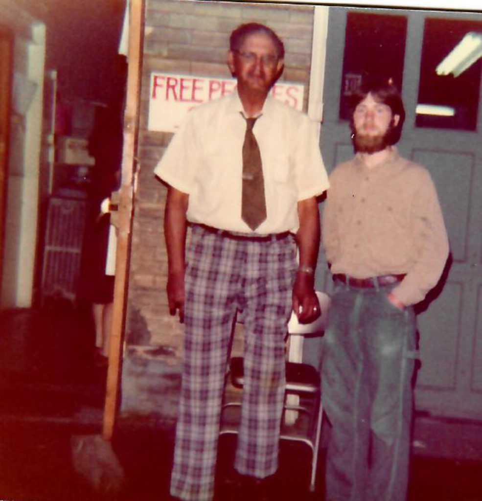Lesley Riddle in check pants and a tie standing beside a shorter bearded man in front of the Genesee Co-op Teahouse.