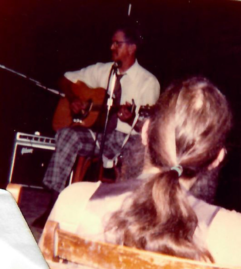 Lesley Riddle on stage with his guitar; a man with a long brown ponytail sits in front of the stage in the audience.
