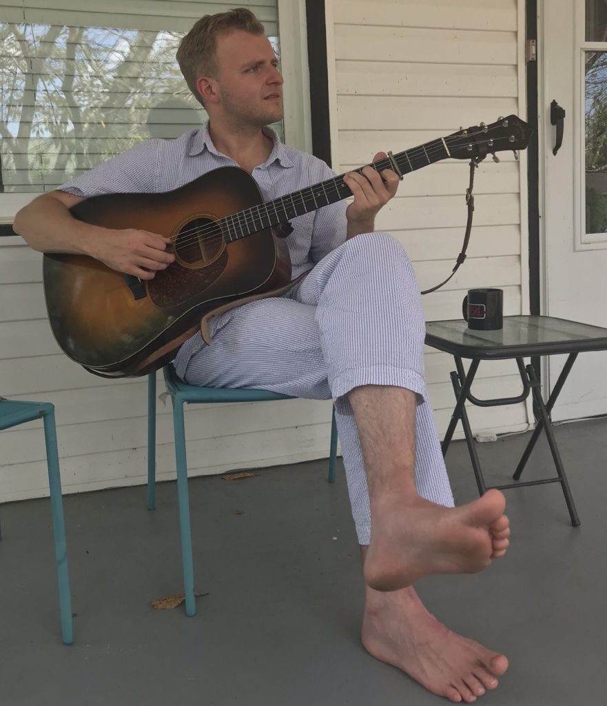 Nathan sitting on the porch, playing guitar with a cup of coffee beside him. He is wearing his pajamas!
