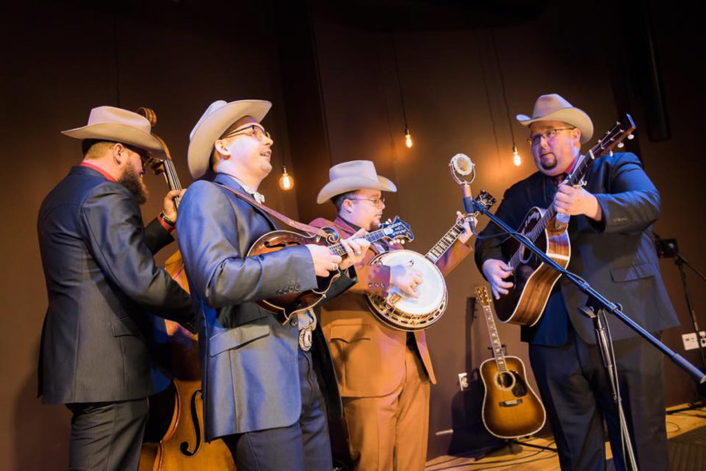 The Po Ramblin' Boys, all wearing suits and hats, gather and play around the mic on stage in the museum's Performance Theater.