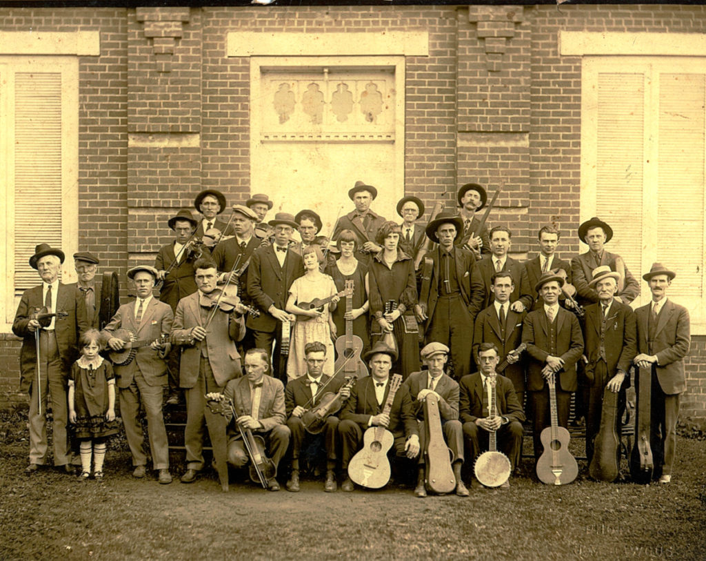 Large group of musicians standing with their instruments in front of what looks to be a large church or civic building; the five members of The Powers Family are seen in the center of the group.
