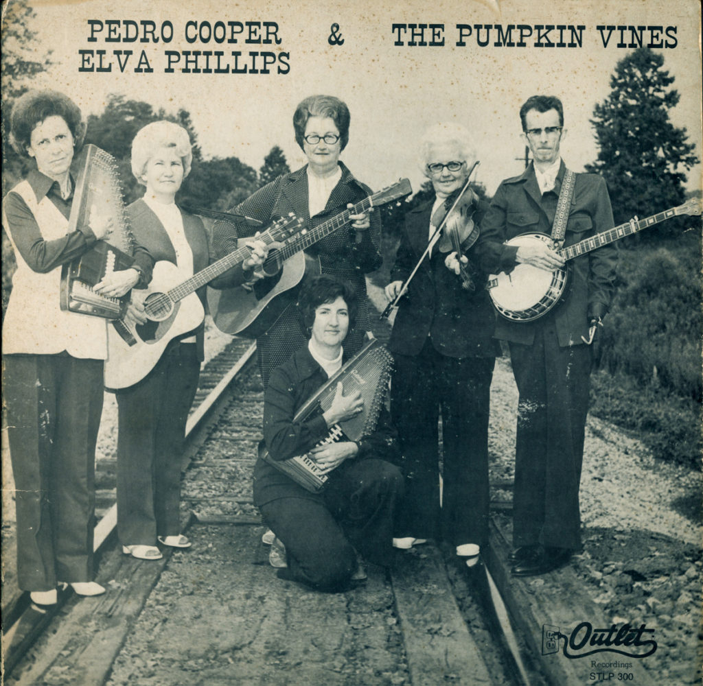 The photograph shows six band members -- five women playing autoharps, guitars, and fiddle, and Pedro on banjo. They are posted standing on a railway line.