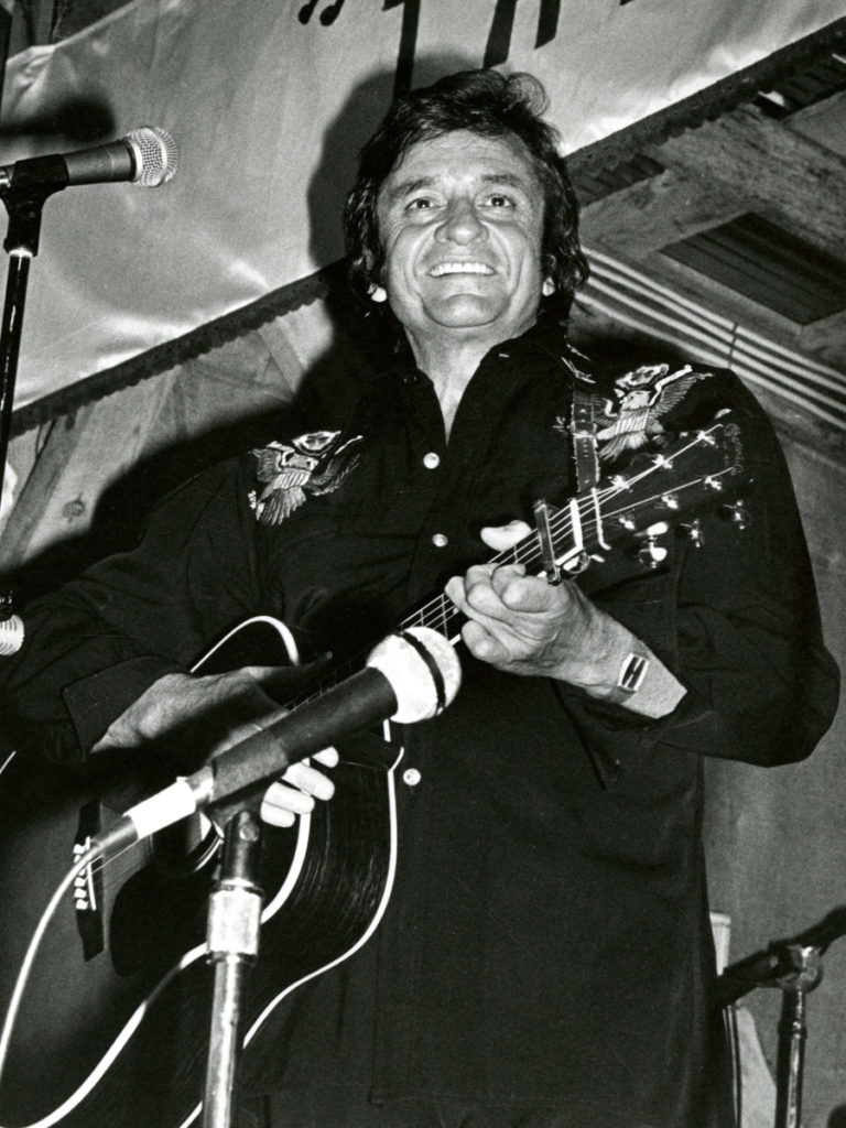 Johnny Cash in a black decorated shirt and holding his guitar on stage in front of a mic; he smiles out at the audience.