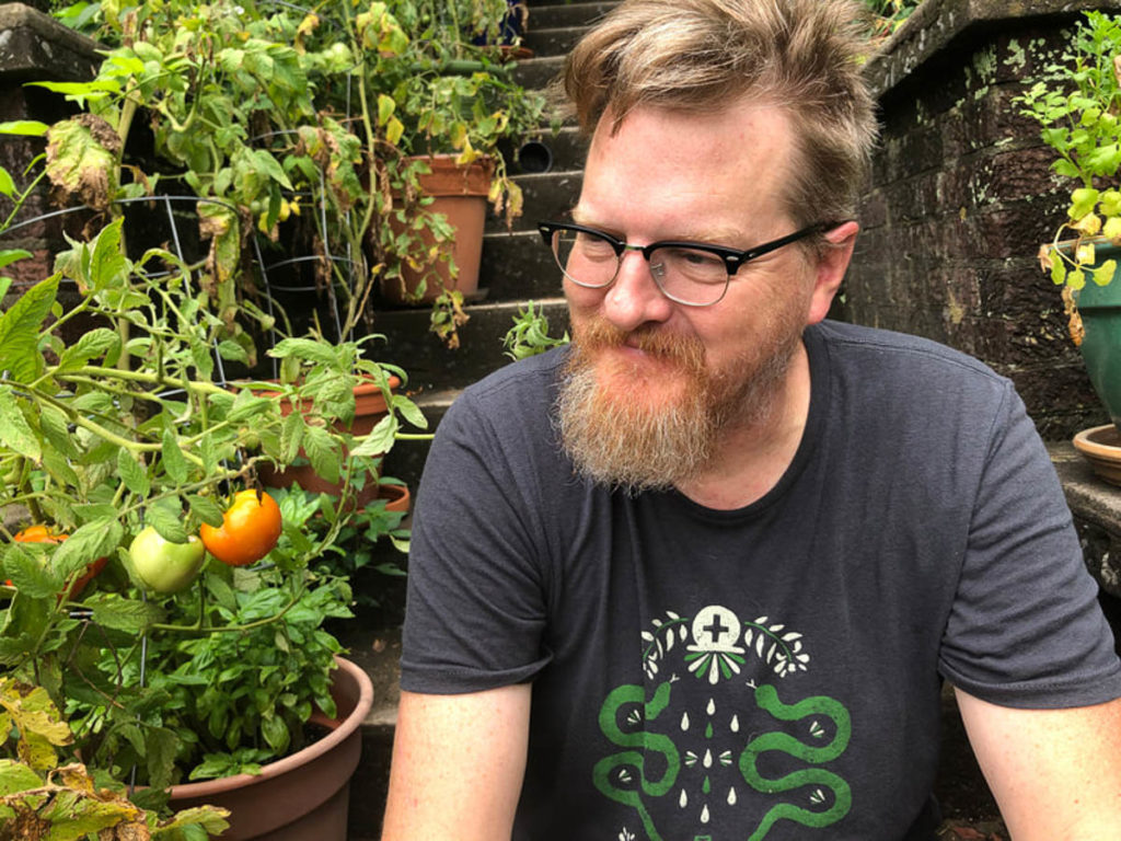 The author is a white man with strawberry blond hair and beard. He is wearing a grey t-shirt with two green snakes in the central design and a pair of black-rimmed glasses. He is seated on some steps surrounded by potted plants, including tomatoes.