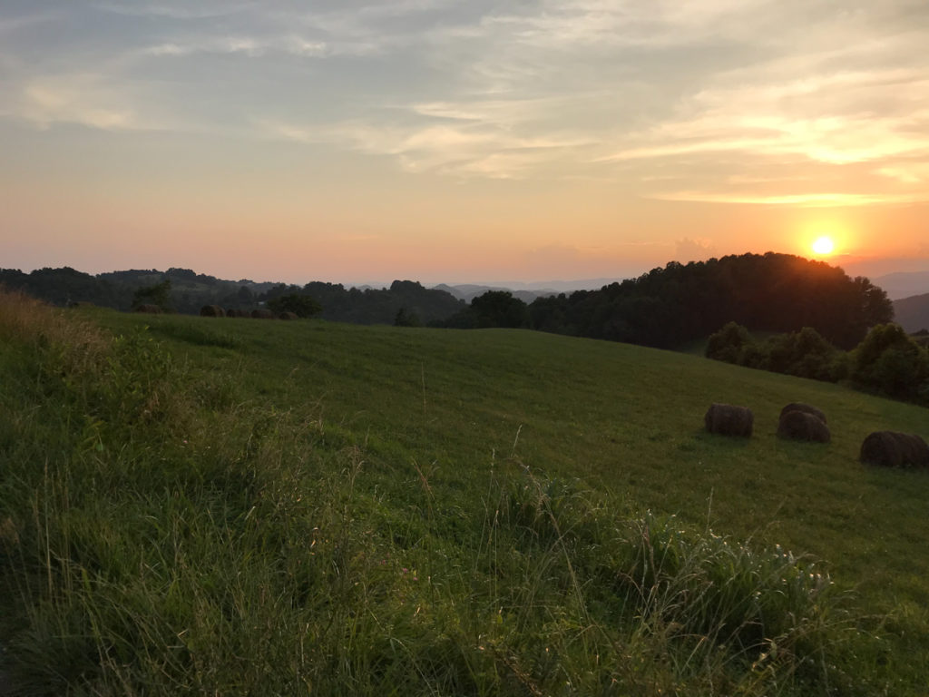 Image of a grassy field and the mountains in the distance with the sun setting behind a hill.