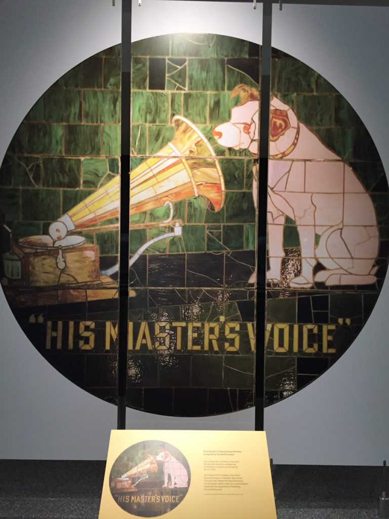 Round stained-glass window, beautiful colors highlighting the design of the Nipper painting and with the word's His Master's Voice beneath the image. A museum label is seen in front of the window.