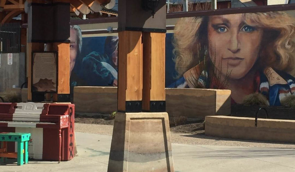 Image of a concreted public space with a painted piano in the foreground and a mural in the background. The mural has a wall-sized portrait of a young Tanya Tucker (head and shoulders, blond wavy hair, small smile, blue eyes, collared shirt); there is the painting of a man to the left on the wall but it is unclear who he is.