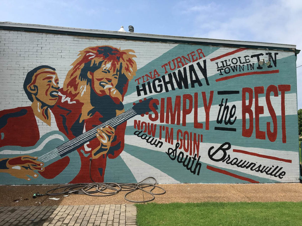An image of a wall mural in a graphic style showing two African-American musicians. The man (Sleepy John Estes) is singing and playing a guitar) and the woman (Tina Turner) is singing and has a big hairstyle. Words from their songs and about Brownsville radiate from their images to the right.