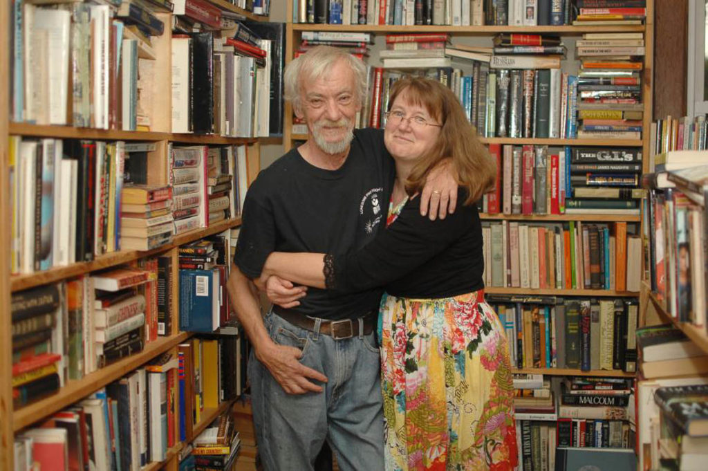 Jack Beck (white-haired with a beard, wearing a black t-shirt and jeans) and Wendy Welch (brown-haired and wearing a black top with a colorful print skirt or dress) stand with their arms around each other in front of floor to ceiling shelves of books.