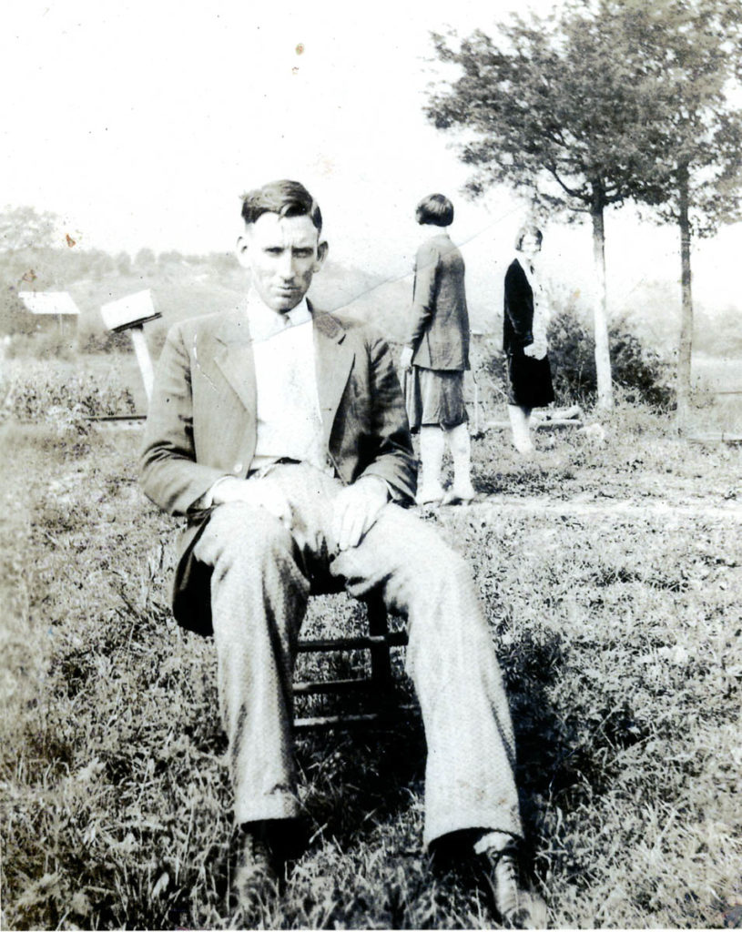 Black-and-white photograph of A. P. Carter sitting in a chair outside, looking straight at the camera. Two women can be seen in the background behind him though they are not the subjects of the picture.
