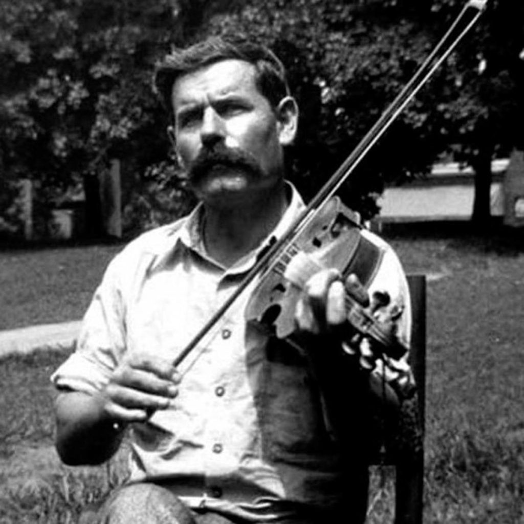 Black-and-white photograph of a dark-haired man seated on a chair outside. He has a large moustache and is holding his fiddle to his shoulder with the bow poised to play.