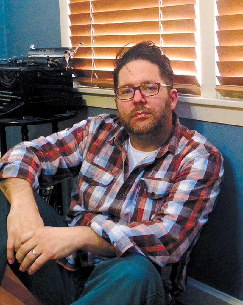 A man with dark brown hair and a short beard sits on the floor in front of a window. He is wearing glasses, a plaid/flannel shirt, and jeans. Beside his is an old typewriter on a table.