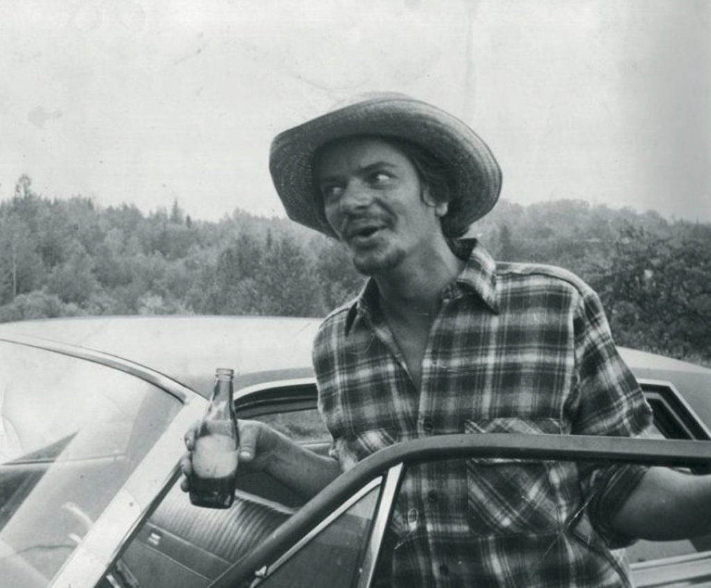 Black-and-white photograph of Michael Hurley in a check shirt and cowboy-style hat, standing beside a car and holding a drink bottle.