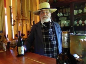 A bearded Michael Henningsen standing behind a bar, holding a bottle of wine. He is wearing period clothing including a straw hat with a black ribbon, a jacket and plaid vest and a neckerchief. 